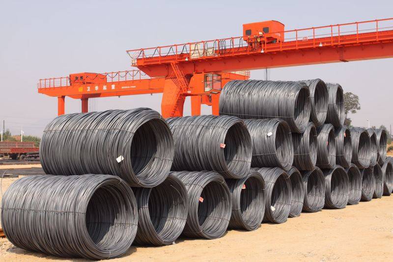 Coils of steel wire rod being loaded into a large cargo ship at a busy port with a crane visible.