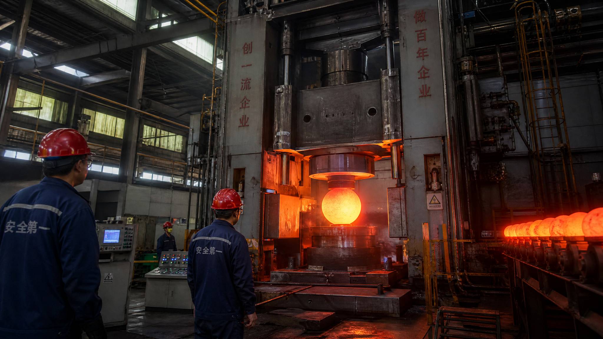 A large automated forging press shaping red-hot steel into spheres inside a modern Chinese factory, with workers monitoring.