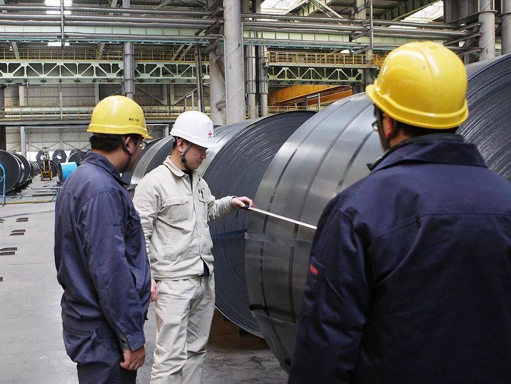 Diverse team of engineers with Asian Chinese faces in safety gear, standing in a modern steel factory with GI and PPGI coils.